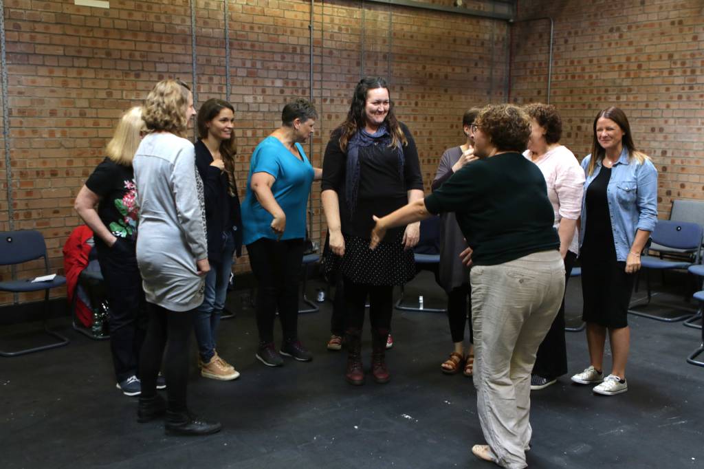 Image of women standing around in a group in a workshop space with a black floor and brick walls. They seem to be in the middle of an exercise, listing to someone as she gesticulates.